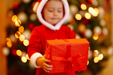 Fototapeta na wymiar Christmas gift in the hand of Little child. Kid in santa costume holding red present box on the background of Christmas lights and tree. Winter holiday, New Year.