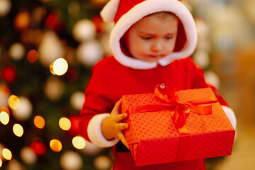 Fototapeta na wymiar Christmas gift in the hand of Little child. Kid in santa costume holding red present box on the background of Christmas lights and tree. Winter holiday, New Year.