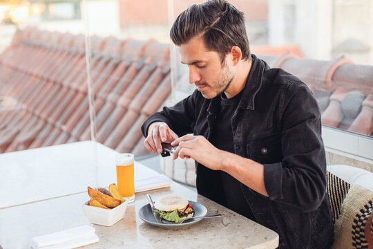 Young blogger man taking pictures of his food in a rooftop restaurant. A Burger, beer and french fries on the table 
