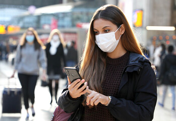 Student girl using mobile phone wearing protective face mask in airport or train station with blurred people on the city background