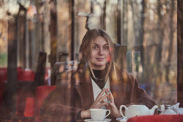 Slightly blurry portrait of a charming girl, made through the window of a cafe. The window reflects the autumn Park.