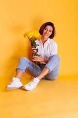 Studio shot on yellow background. Happy caucasian woman short hair wearing casual clothes, white shirt and denim pants, holding bouquet of yellow asters.  