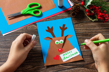 Instruction step 8. Merry greeting card with a Christmas deer on a wooden table. Handmade. Children's creativity project, crafts, crafts for kids.