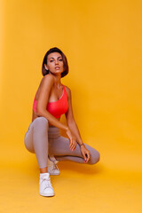 Sport and yoga lifestyle concept. Young fit pretty strong woman dressed in sport clothes, stylish top and leggings, poses against yellow background. Studio shot.   