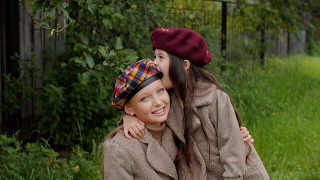 Cute happy little girls in berets hugging outdoors. Adorable cheerful little sisters in coats and hats having fun together and embracing on green lawn in garden