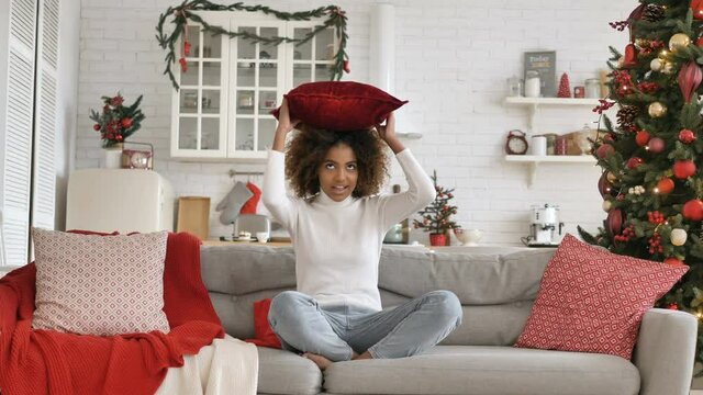 Cheerful kinky haired lady with dark skin takes pillow puts on head and sits in lotus pose on sofa near Christmas tree slow motion