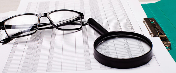 On the table are black-framed glasses, reports and a magnifying glass. Business concept