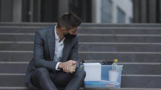Unemployed businessman lost his business. Anxious concept. Workless man in despair. Fired male office worker in medical mask sitting on stairs in depression with box of stuff