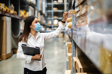 woman worker with medical mask holding clipboard and checking inventory in warehouse during...