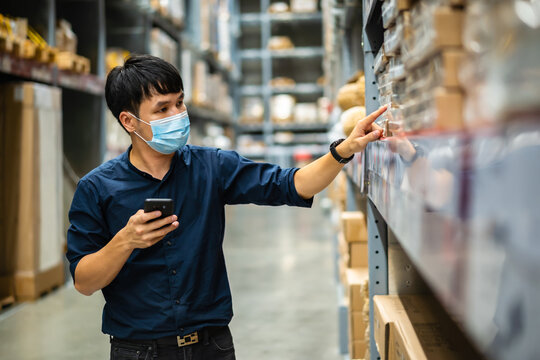 man worker with medical mask using smartphone to checking inventory in warehouse during coronavirus (covid-19) pandemic