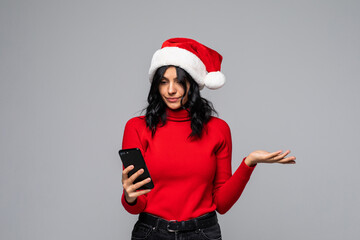 Portrait of a confused cute woman in dress talk at mobile phone. emotional girl in santa claus christmas hat isolated on gray background.