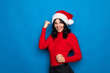 Young beautiful woman wearing a christmas hat over blue background very happy and excited making winner gesture with raised arms, smiling and screaming for success.