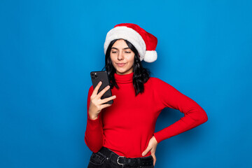 Beautiful woman typing on the phone with christmas hat is posing isolated on blue background.