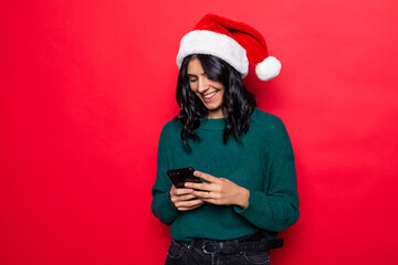 Young woman wearing santa hat typing phone isolated on red background