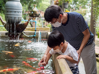 Asian family travel outdoor together with wearing face mask protecting from covid-19. Cute child and father feeding milk to fish. Travel and health care concept.