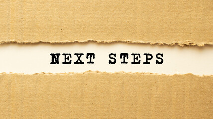Text NEXT STEPS appearing behind torn brown paper. Top view.