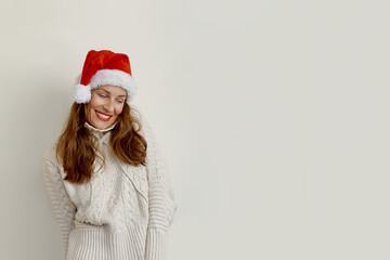 The average adult beautiful woman wearing a sweater and a Santa Claus hat stands on a beige background, looking down and smiling