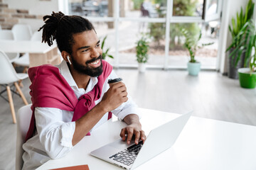 Smiling african american guy drinking coffee while working with laptop - Powered by Adobe