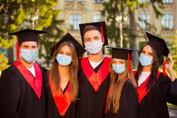 Photo portrait of five graduates in robes and hats with tassel wearing face masks and hugging each...