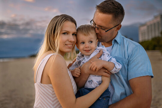 portrait of happy caucasian family. Mother and father holding a little son in arms. Dad kissing a boy. Summer sunset at the beach. Image with selective focus.