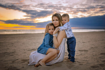 Fototapeta na wymiar happy beautiful mother with her daughter sitting on her laps and baby son hugging her on the beach in sunset. Beautiful cloudy sky on background. Image with selective focus