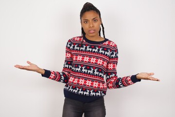 Young beautiful African American woman wearing Christmas sweater against white wall looks uncertain...