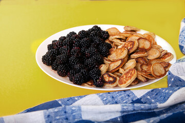 Minipancakes with blackberries on a skewer, two forks on a blue napkin on a yellow background. Dessert.