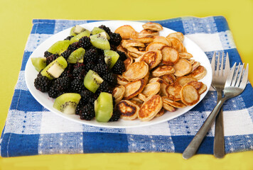 Minipancakes with blackberries and kiwi slices on a plate, two forks on a blue linen napkin on a yellow background. Dessert.