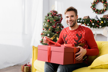 Obraz na płótnie Canvas happy man in red sweater holding wrapped present while sitting on sofa with blurred christmas tree on background