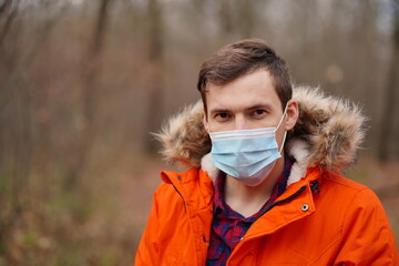 Portrait of adult man in medical mask in autumn forest. Close up of male in orange jacket and protective mask on walk. Concept of prevention from viral infection.