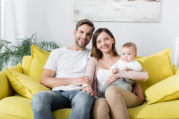 Fototapeta premium cheerful parents smiling near baby boy while sitting on sofa
