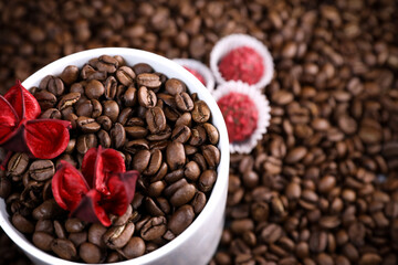 White cup full of coffee beans on a coffee beans background with red chocolate candy and red flowers. Morning espresso. Coffee mug.