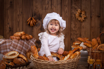 Happy baby in the image of a chef, soiled in flour, laughing merrily kneading the dough. playing the role of a chef, bakery, a lot of bread rolls butter pretzels on a wooden background hanging bagels