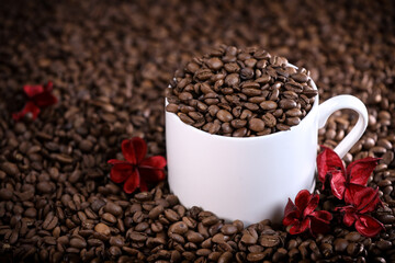 White cup full of coffee beans on a coffee beans background with red flowers. Morning espresso. Coffee mug.