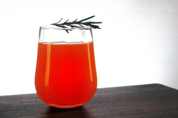 Semicircular glass of fresh orange juice with rosemary sprig on top staying on a wooden table against light background