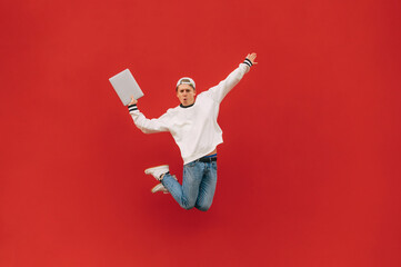 shocked guy in stylish casual clothes jumps on a background of a red wall with surprise on his face and a laptop in his hands. Background. Isolated.