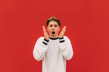 Shocked young man isolated on red background, looks at the camera with his hands raised to his face and looks at the camera with a surprised face