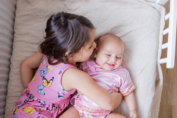 Happy little girl lying down on a bed and hugging her smiling baby sister