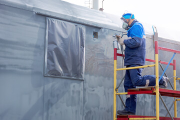 Industrial work. Priming of metal products from the compressor gun. A worker in overalls and a respirator paints the body of a truck trailer or a metal car.