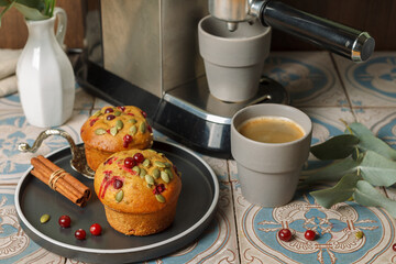 Two muffins with berries and seeds on a black plate and a coffee machine pouring espresso into gray cups.