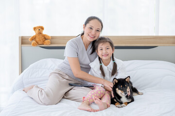 Asian mother and girl sit on white bed with black Shiba dog and look to camera with smiling in bedroom. Concept of happy family feel relax to stay home with their own pets