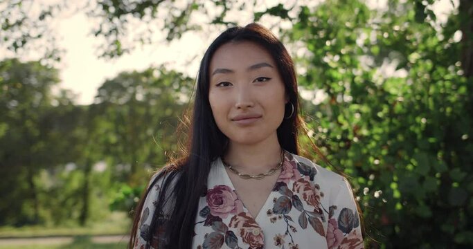 Portrait of asian female person with dark long hair looking to camera in daylight. Crop view of serious young woman posing and smiling while standing in park . Concept of lifestyle