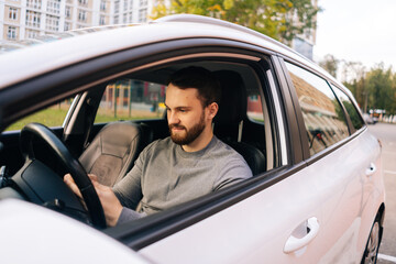 Fototapeta na wymiar Confident bearded young man sitting in car and typing online message on cell phone, side view. Handsome businessman using mobile phone in auto. Male sitting in modern vehicle and works on smartphone.