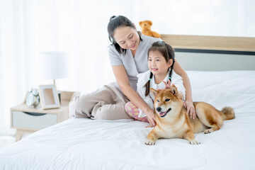 Asian mother and girl sit on white bed with black Shiba dog and look to left side with smiling in bedroom. Concept of happy family feel relax to stay home with their own pets.