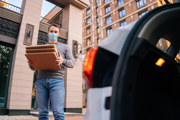 Low-angle shot of delivery man wearing medical protective mask standing at city street with modern building near trunk of car and holding carton boxes with hot pizza. Concept of food delivery service.