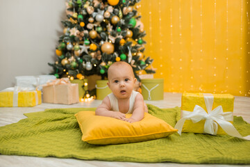 a baby boy in a white bodysuit lies on a green knitted blanket with a gift on the background of a Christmas tree