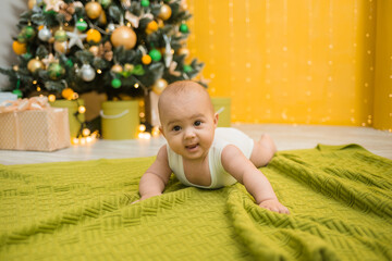 a cheerful little boy in a white bodysuit lies on a green knitted blanket against the background of a Christmas tree