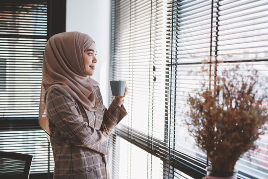 Young asian muslim business woman in smart casual wear standing beside the glass wall her hands holding a cup of coffee and looking outside at the modern office.