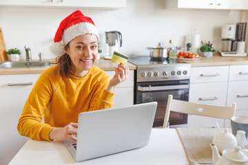 Woman in Santa hat shopping online and paying with gold credit card. Young girl with laptop buying Christmas gift present on Internet on kitchen indoor. Online Christmas shopping concept.