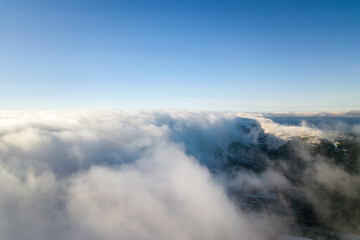 Aerial view from above of white puffy clouds covering snowy mountain tops in bright sunny day.